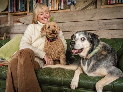 Blonde woman in white jumper sits on couch smiling at her two dogs