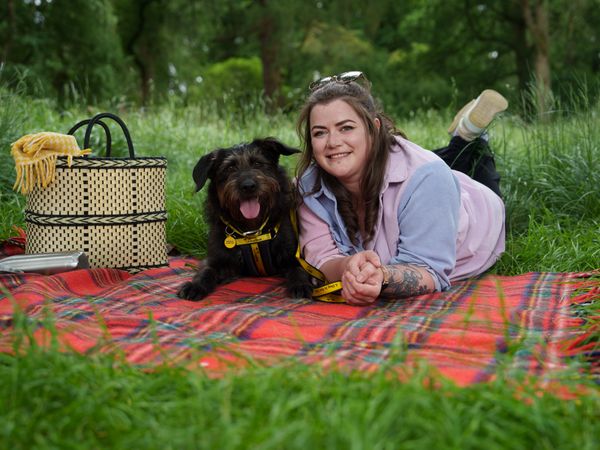 A woman and a black and brown longhaired dog lie on a picnic blanket in a park