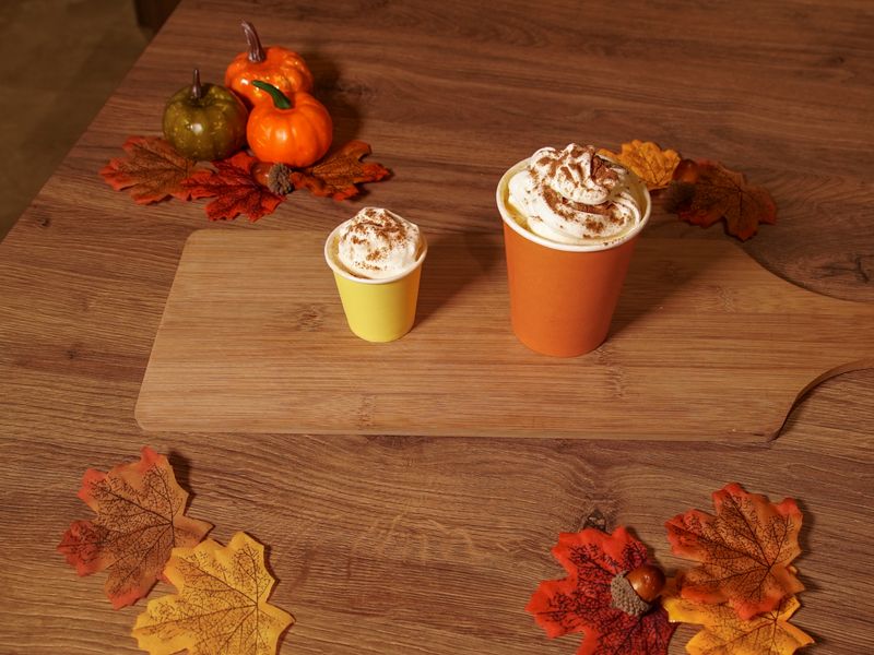 Photo of some pumpkin spiced pup-cups, one in a small yellow cup and a larger one in an orange cup. Autumn leaves in red, orange and yellow and small pumpkins, decorate the table.