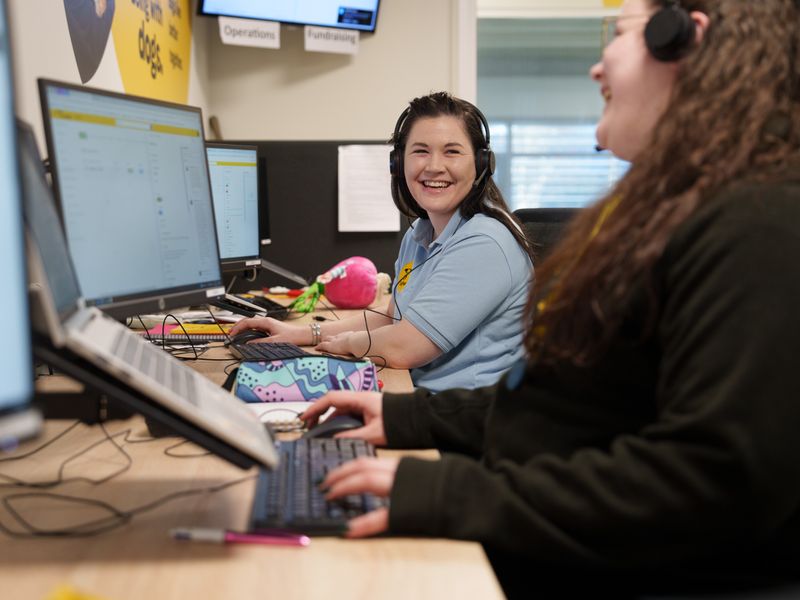 Two women sitting at computers in an office with headsets on, they ae looking at each other an smiling